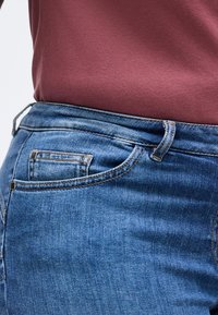 Κοντινό πλάνο τσέπης τζιν μπλε και θηλιάς ζώνης με πουκάμισο μπορντό που είναι μπαλωμένο στη μέση.