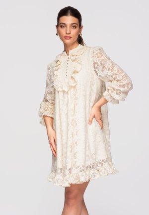 Femme portant une robe en dentelle ivoire à manches longues avec des volants le long du col, de la poitrine, des poignets et de l'ourlet, debout avec la main sur la hanche devant un fond uni.