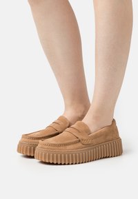 Mocasines de ante marrón con un diseño de parte superior acolchada y suela de goma ribeteada. Presenta un sutil acento de costura a lo largo de la puntera.