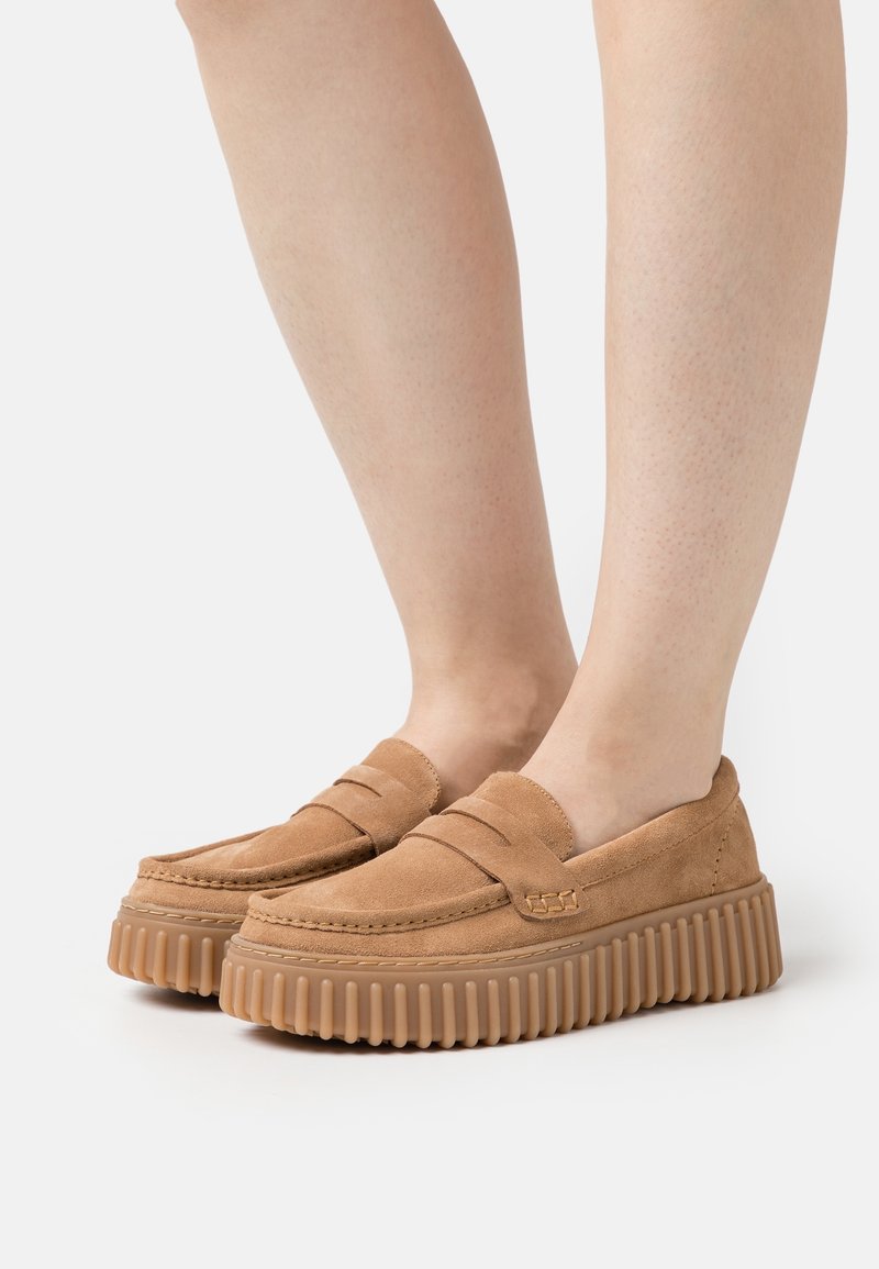 Mocasines de ante marrón con un diseño de parte superior acolchada y suela de goma ribeteada. Presenta un sutil acento de costura a lo largo de la puntera.