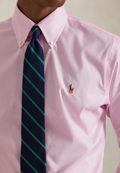 Muž nosí světle růžovou košili s knoflíky a tmavě modro-zelenou pruhovanou kravatu s malým vyšívaným hráčem póla na koni.