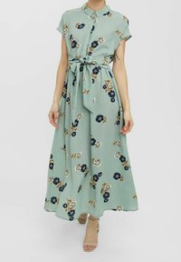 Blommig maxiklänning i ljusgrön tyg med korta ärmar, knäppning med knappar, midjeband och spridda mörkblå och vita blommönster.
