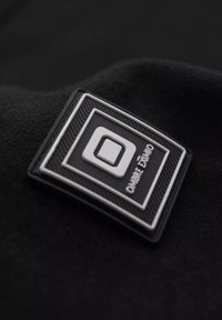 Černá tkanina má obdélníkovou gumovou nášivku s texturovaným okrajem. Nášivka zobrazuje slovo "OMBRE" a čtvercové logo v bílé barvě.