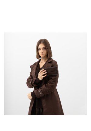 Donna con capelli castani lisci indossa un cappotto marrone scuro e tiene in mano un portafoglio marrone coordinato su uno sfondo bianco semplice.