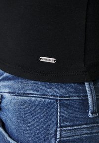 Schwarzes Baumwoll-T-Shirt mit einem kleinen silbernen "HOLLISTER"-Label, kombiniert mit hellblauen Jeans mit sichtbaren Nahtdetails.