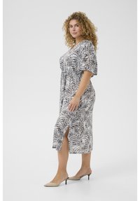 Robe fluide de longueur genou en tissu léger avec des motifs de feuilles en bleu marine et blanc, dotée de manches courtes et d'une fente à l'avant. Associée à des talons beiges.