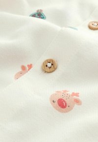 Baumwollstoff mit einem cremefarbenen Hintergrund, der ein Muster aus bunten Rentieren und Bäumen zeigt. Holzknopf mit floraler Gravur.