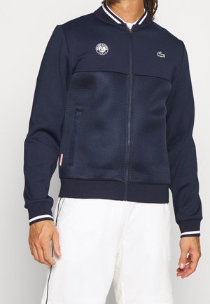 Mand iført en mørkeblå lynlåstrøje med Roland Garros og Lacoste logoer, kombineret med hvide shorts mod en ensfarvet baggrund.