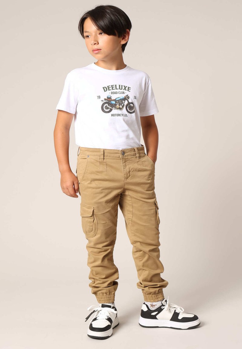 Khaki cargobroek met elastische boorden, twee zijzakken en een klassiek wit T-shirt met een motorfietsafbeelding. Zwart-witte sneakers.