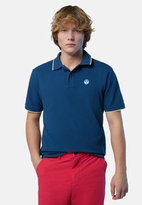 Polo shirt blu navy con colletto giallo a contrasto e logo sul lato sinistro del petto. Indossato con pantaloni corti rossi vivaci, mostrando una vestibilità rilassata.