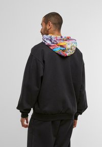 Mann mit dem Rücken zur Kamera, der einen schwarzen sweatshirt mit buntem, gemustertem Kapuzenshirt und passenden schwarzen Hosen trägt, vor einem schlichten Hintergrund.