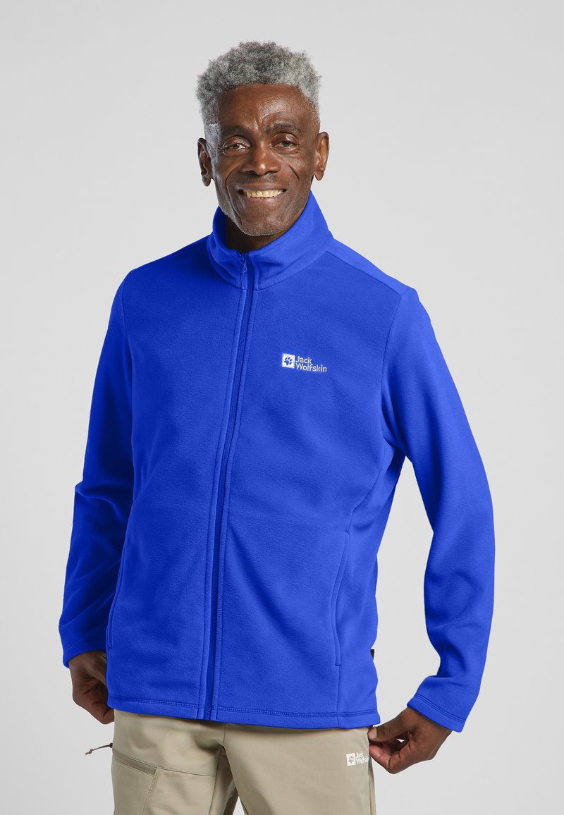 Blauwe fleecejas met hoge kraag, volledige rits en zijzakken. Bevat een logo op de linkerborst. Glad textuur.