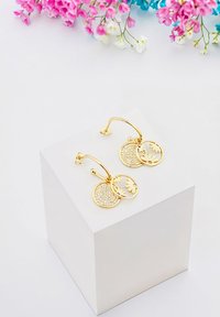 Guldörhängen med hoops har två cirkulära berlocker: en med en texturerad yta och den andra med blomsterutsnitt, utställda på ett vitt podium.