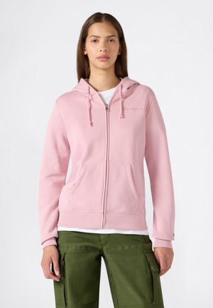 Mujer joven con sudadera rosa claro con cremallera y bolsillos delanteros y pantalones cargo verde oliva sobre un fondo blanco liso.
