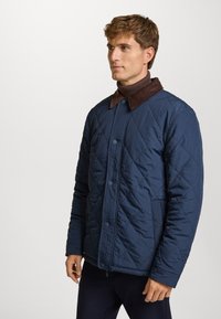 Chaqueta acolchada azul marino con un cuello de pana marrón, que cuenta con botones de presión y mangas largas. Textura suave y patrón de costura en diamante.