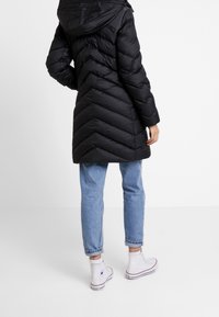 Schwarzer Steppmantel mit Chevron-Muster, Kapuze und langem Schnitt, kombiniert mit hellblauen hochgekrempelten Jeans und weißen High-Top-Sneakers.