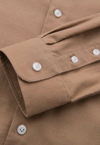 Poignet de chemise marron à manches longues avec boutons blancs, couture visible et tissu texturé en gros plan.