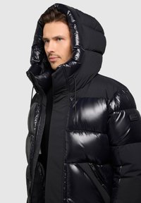 Čierna prešívaná bunda s lesklým prešívaným dizajnom, veľkou kapucňou a zapínaním na zips. Vyrobená z izolovaného, hladkého materiálu.