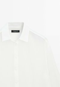 Camicia bianca con colletto classico e chiusura con bottoni. Realizzata in tessuto morbido con una lucentezza sottile, presenta un'etichetta del marchio sul colletto.