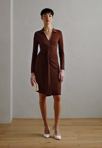 Robe longue marron à manches longues avec décolleté en V et devant froncé, présentant une silhouette ajustée. Des escarpins blancs et une petite pochette complètent le look.