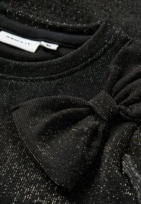 Czarna tekstylna bluzka z metalicznym połyskiem, z okrągłym dekoltem i ozdobnym fasonem w postaci kokardy na przodzie.