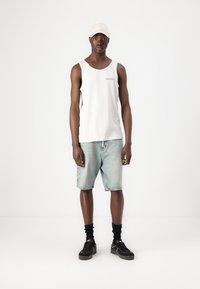 Canotta bianca, pantaloni shorts di denim azzurro chiaro, sneakers nere con dettagli bianchi e un cappellino beige. Design semplice e fit casual.