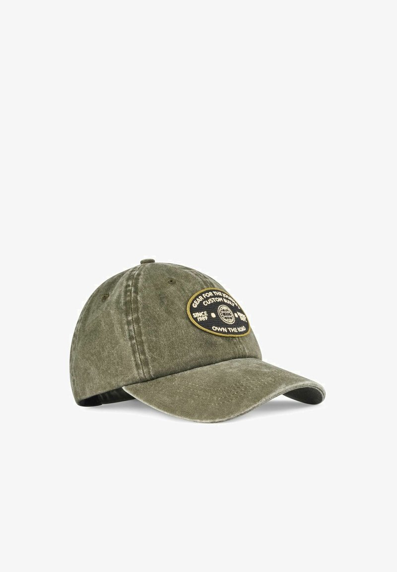 Oliwkowa bawełniana czapka z zakrzywionym daszkiem, z okrągłą czarno-złotą naszywką na przodzie. Napis brzmi: „Sprzęt na długą drogę.”