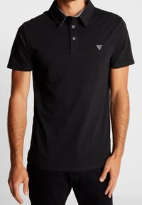 Hombre vistiendo una camiseta polo de manga corta negra con dos botones y un pequeño logo triangular en el lado izquierdo del pecho.