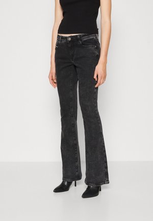 Kvinde iført falmede sorte bootcut-jeans, sort ærmeløs top og sorte spidse hæle støvler står mod en ensfarvet lys baggrund.