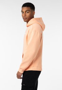 Pfirsichfarbener Hoodie aus weichem Stoff mit Kängurutasche, Kordelzugkapuze und gerippten Bündchen. Mit weißem Text auf der Vorderseite.