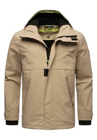 Beige waterdichte jas met capuchon, voorzien van een zwarte fleece voering en groene accenten. Inclusief ritszakken aan de voorkant en op de borst.