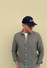Grijze linnen shirt met knopen aan de voorkant, opgerolde mouwen; zwarte pet met de tekst "Bonded"; zonnebril; effen achtergrond.