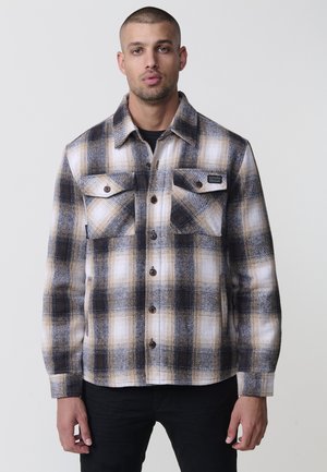 Camisa de franela a cuadros en crema, marrón y negro. Presenta dos bolsillos en el pecho, cierre de botones y un corte regular con mangas largas.