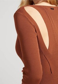 Gros plan d'une personne portant un pull en maille côtelée de couleur rouille avec de grandes découpes aux épaules et au haut du dos.