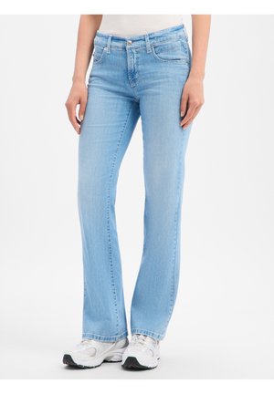 Femme portant un jean bleu clair droit avec des baskets blanches, debout sur un fond blanc uni.