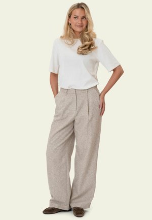 Hosszú szőke hajú nő fehér rövid ujjú felsőben, bézs bő szárú nadrágban és barna mokaszinban, kezét a zsebében tartva áll.