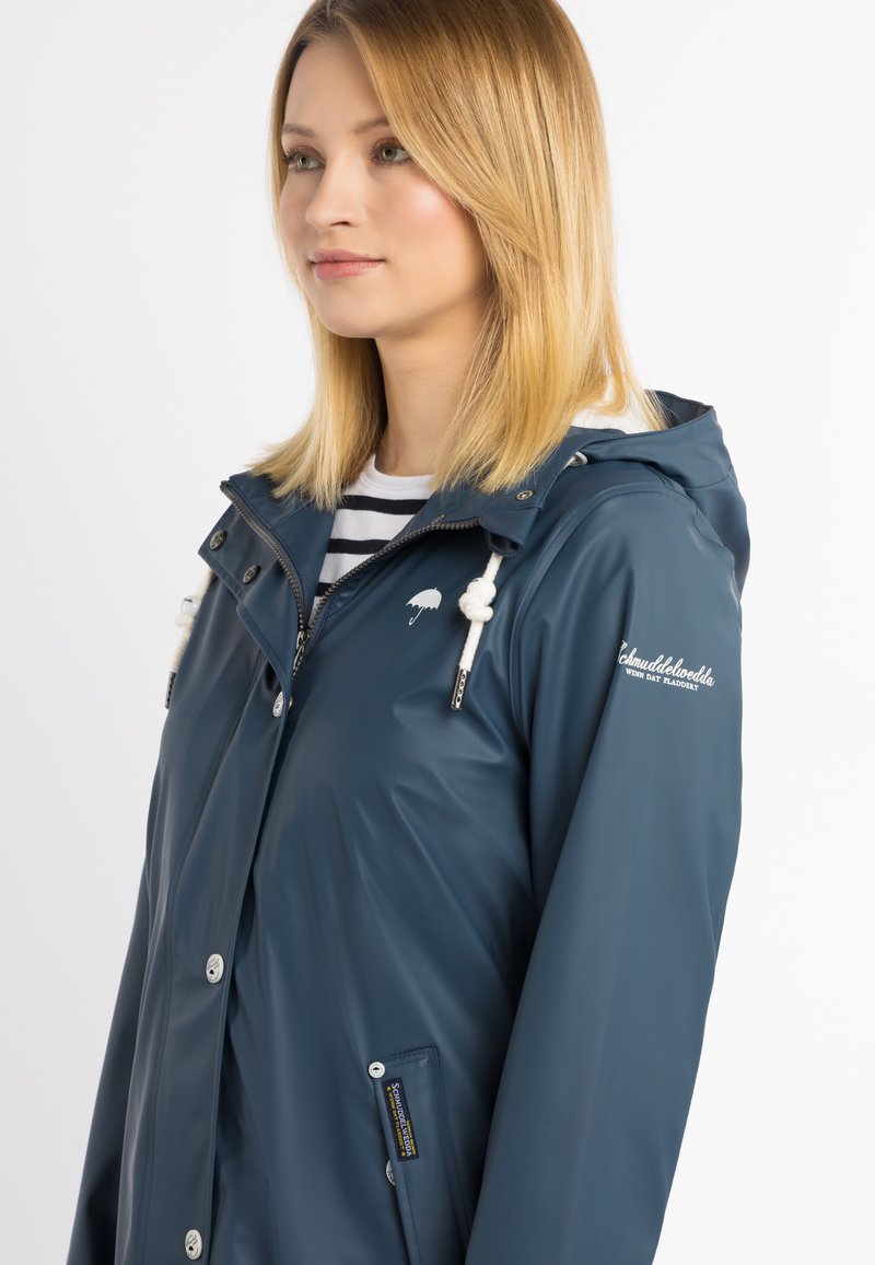 Chaqueta impermeable azul con capucha, cremallera frontal, cordones blancos y detalle de logo en la manga. Textura suave, cierres de botón.