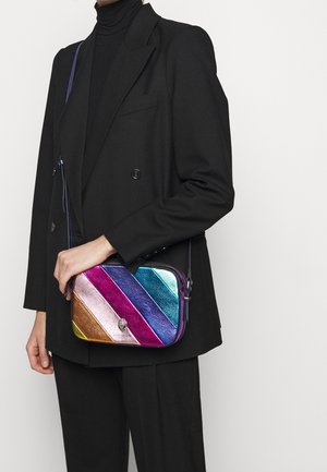 Personen iført en sort jakkesæt og turtleneck holder en lille rektangulær crossbody taske med skinnende diagonale metalliske striber i guld, pink, lilla, blå og teal.