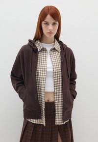 Sudadera marrón con cordón, superpuesta a una camisa de cuadros en beige y marrón, combinada con una falda de cuadros con una abertura alta.
