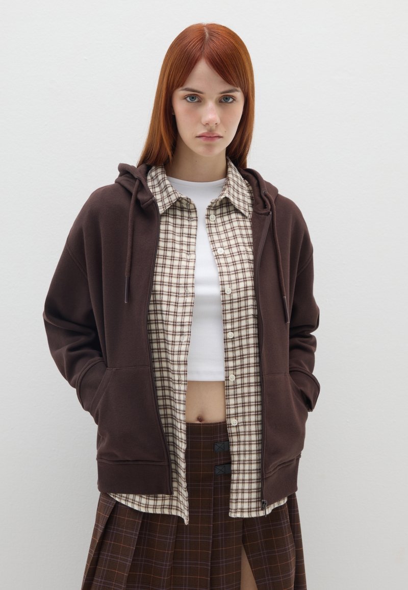Sudadera marrón con cordón, superpuesta a una camisa de cuadros en beige y marrón, combinada con una falda de cuadros con una abertura alta.