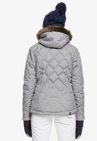 Grauer gesteppter Mantel mit Kapuze und Kunstfellbesatz, mit einer taillierten Passform und sichtbaren Nähmustern. Kombiniert mit einer navy blauen Strickmütze und Handschuhen.