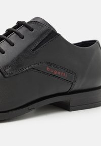 Chaussure en cuir noir à texture lisse, dotée de lacets et d'un logo discret rouge. Talon bas et design épuré avec une couture contrastante.