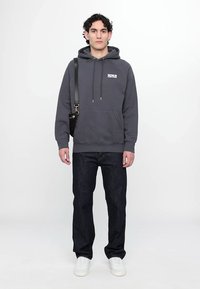 Grijze hoodie met een buidel en koordjes, gecombineerd met donkere spijkerbroeken en witte sneakers, wat een casual, gelaagde uitstraling creëert.