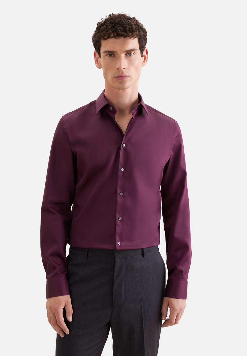 Homme aux cheveux bouclés foncés portant une chemise longue à manches longues violet foncé boutonnée et un pantalon gris foncé sur fond blanc.