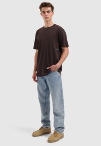 Young man standing with one hand in pocket, wearing dark brown T-shirt, light blue jeans, and tan lace-up shoes.