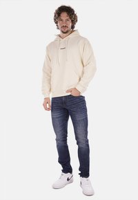 Felpa beige con tasca anteriore, cappuccio e testo del logo nero; abbinata a jeans blu scuro e sneakers bianche. L'uomo è in piedi di fronte alla fotocamera.