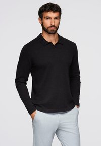 Polo a maniche lunghe nera con colletto classico e vestibilità comoda, abbinato a pantaloni grigi chiari. Tessuto di consistenza morbida.
