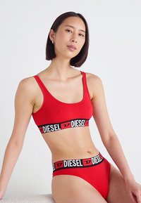 Rdeča bombažna bralette in ujemajoča se bikinka z črnim elastičnim trakom, na katerem je v izmeničnih barvah napis "DIESEL". Gladka tekstura, prilegajoča zasnova.