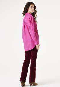 Camisa de botones de color rosa brillante combinada con pantalones de pana burdeos de pierna acampanada. La camisa tiene un corte relajado con un dobladillo curvado y mangas largas.