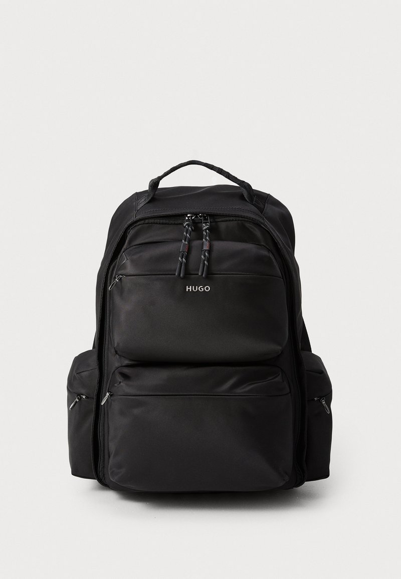 Mochila negra con múltiples compartimentos, hecha de un tejido duradero. Cuenta con cremalleras, un asa con bucle y un logo en la parte frontal.